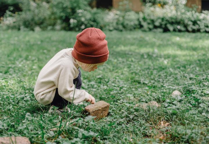 Chłopiec w brązowej kurtce i czerwonej czapce siedzi na trawniku dotykając małego drewnianego elementu